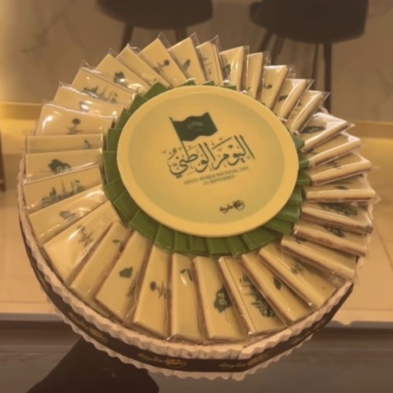 صينية اليوم الوطني بقطع شوكولاتة مغلّفة بالأبيض والأخضر حول قرص بعبارة «اليوم الوطني»
