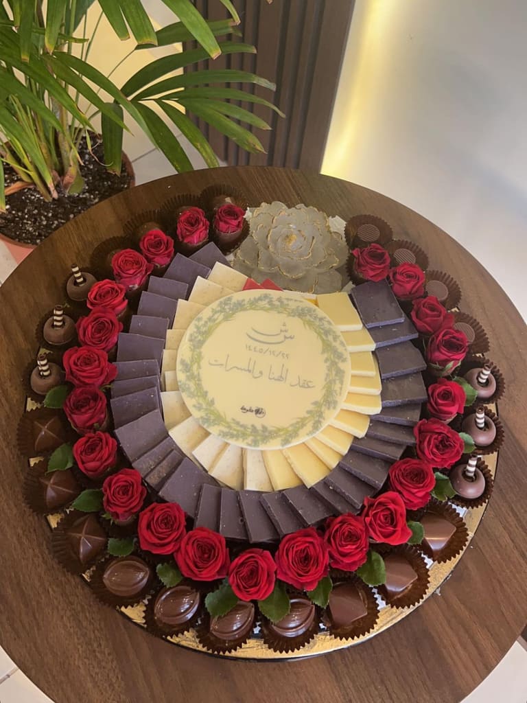صينية عقد قران بحلقة ورد جوري أحمر حول مراوح من الشوكولاتة البيضاء والداكنة، وفي وسطها قرص بعبارة «عقد الهنا والمسرات» وتاريخ هجري